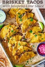 An overhead view of a sheet pan filled with two rows of baked brisket tacos with a colorful bowl filled with pickled red onions in one corner and another bowl filled with jalapeño-lime sauce in the opposite corner.