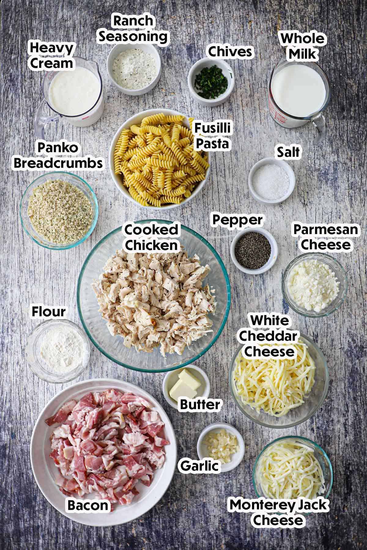 An arrangement of ingredients for chicken bacon ranch casserole on a grey wooden background including cooked chopped chicken, uncooked pasta, shredded cheese, bacon, garlic, flour, cream, breadcrumbs, and seasonings.