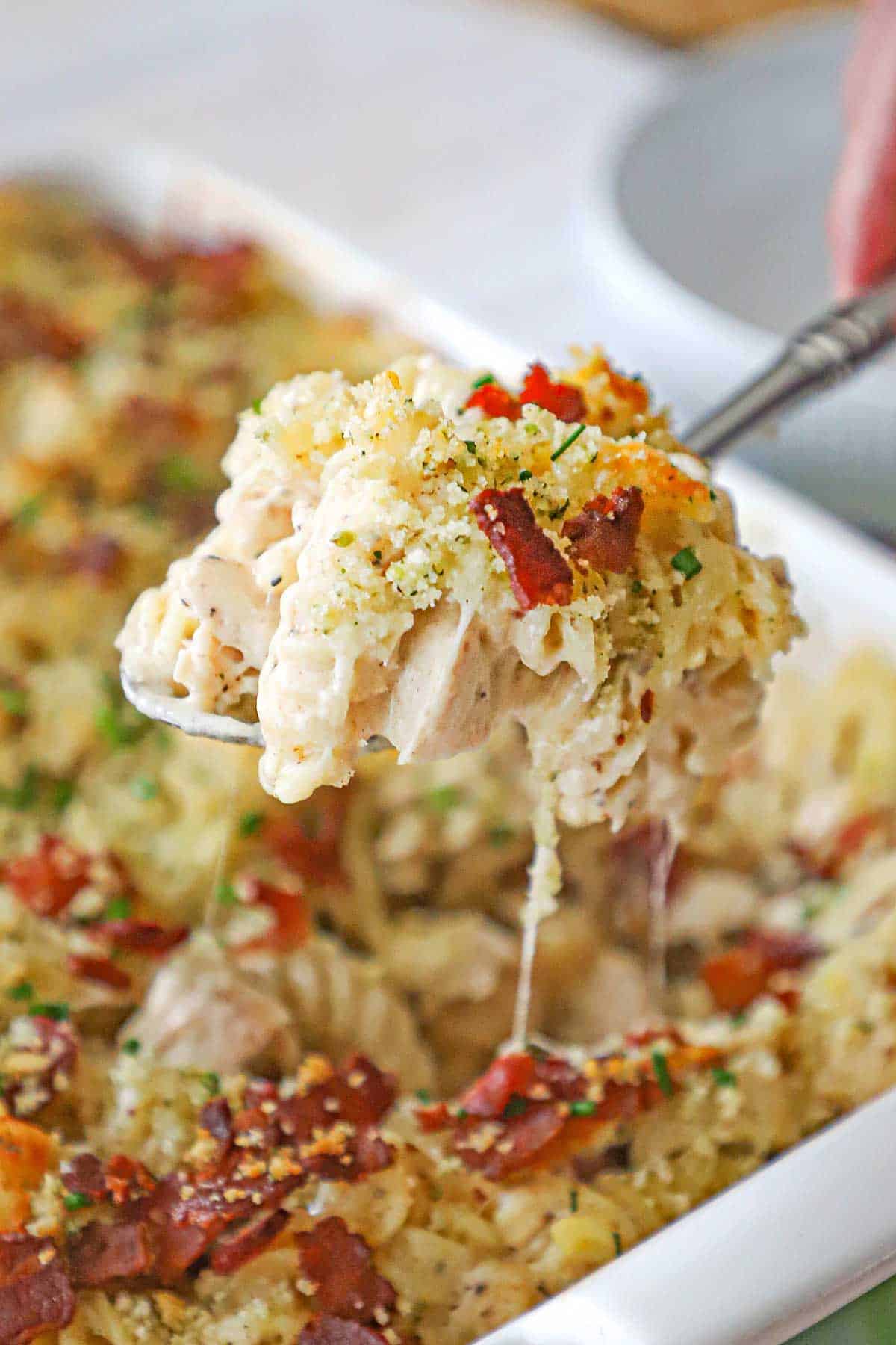 A person using a large serving spoon to lift a serving of chicken bacon ranch casserole from a white baking dish.