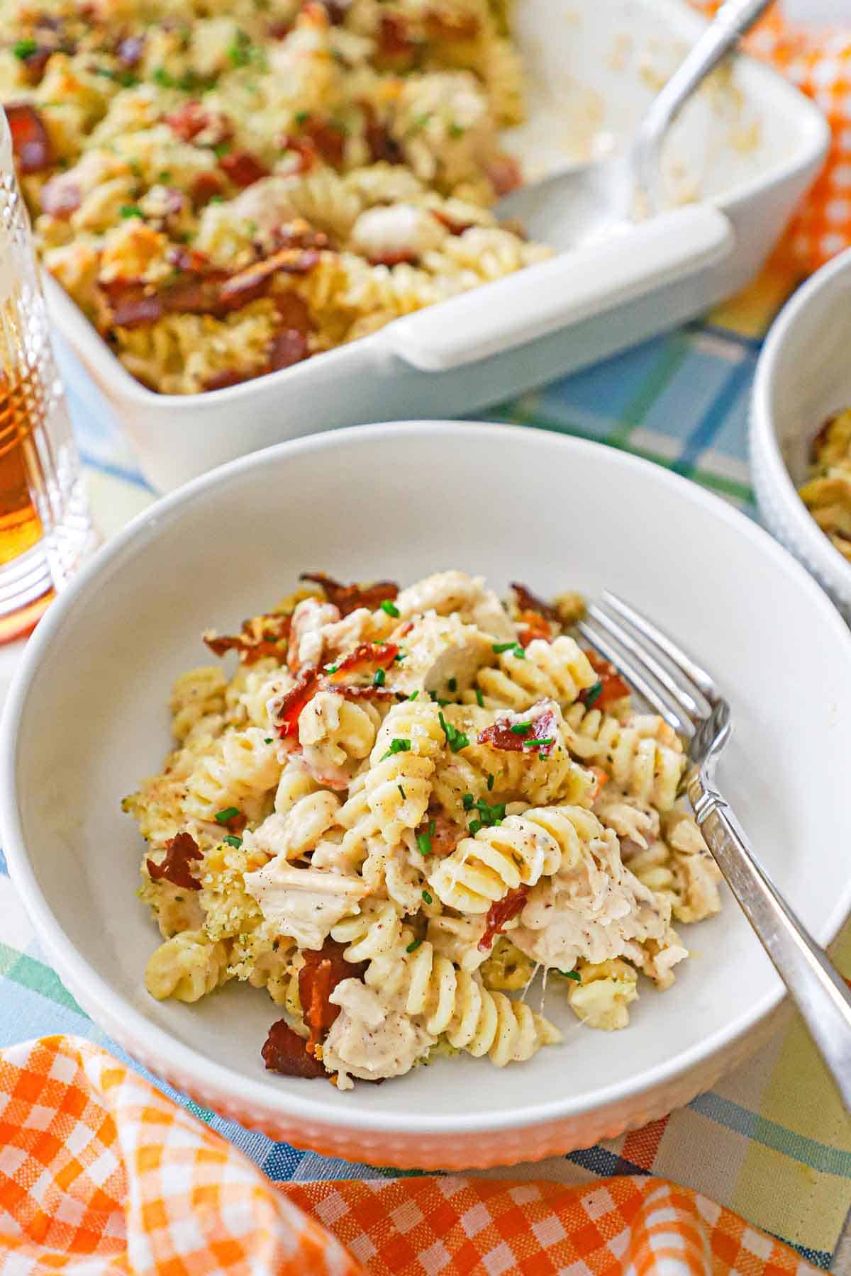 A white pasta bowl filled a serving of creamy chicken bacon ranch casserole with baking dish behind it filled with the same dish.