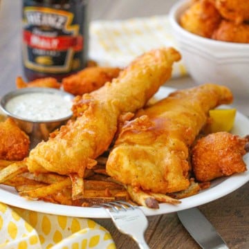 A close-up view of a white dinner plate filled with a serving of Classic Fish and Chips with a condiment holder filled with tartar sauce, a lemon wedge, and Southern hushpuppies nearby.