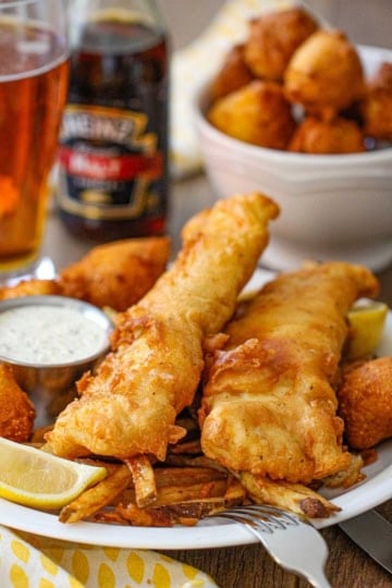 A straight-on view of a white dinner plate filled with a serving of Classic Fish and Chips with a condiment holder filled with tartar sauce, a lemon wedge, and Southern hushpuppies nearby.