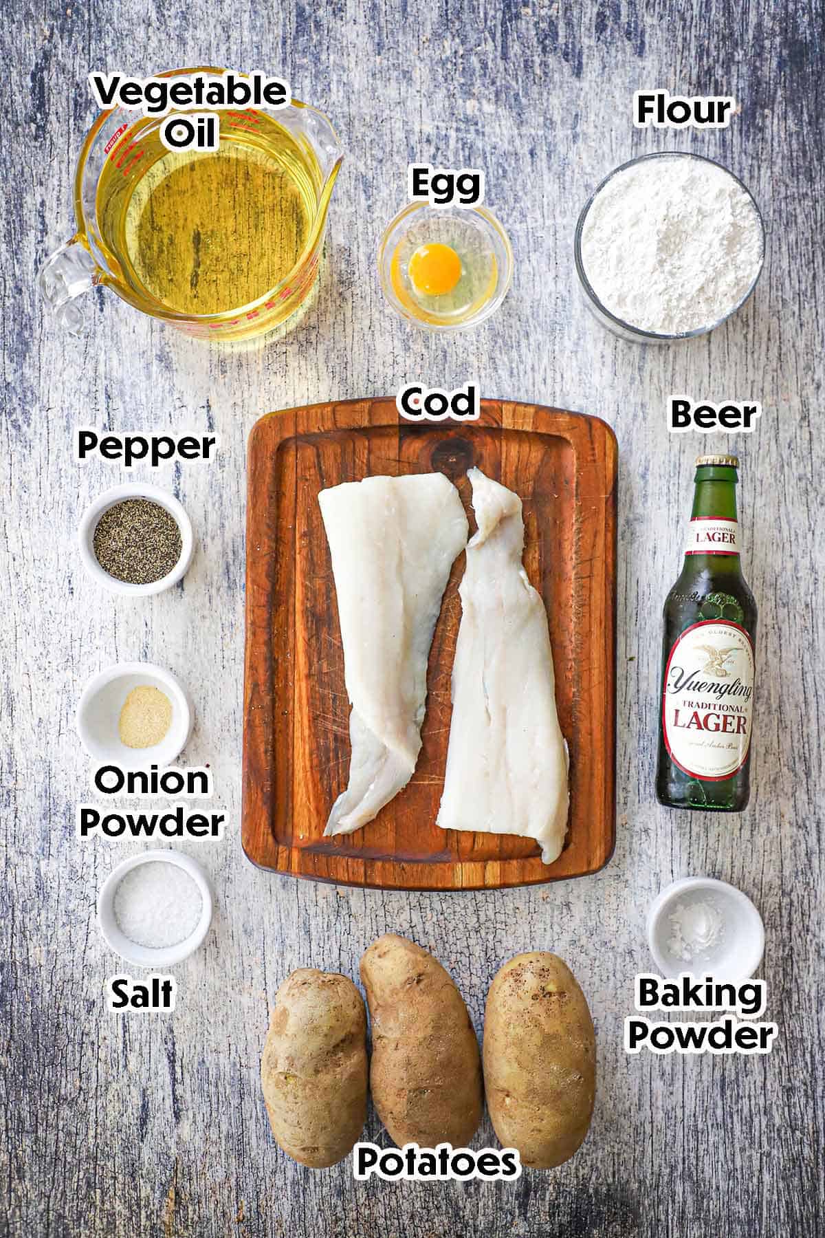 An arrangement of ingredients for classic fish and chips on a grey wooden background including two filets of cod, potatoes, a bottle of beer, flour, egg, oil, baking powder, and seasonings.
