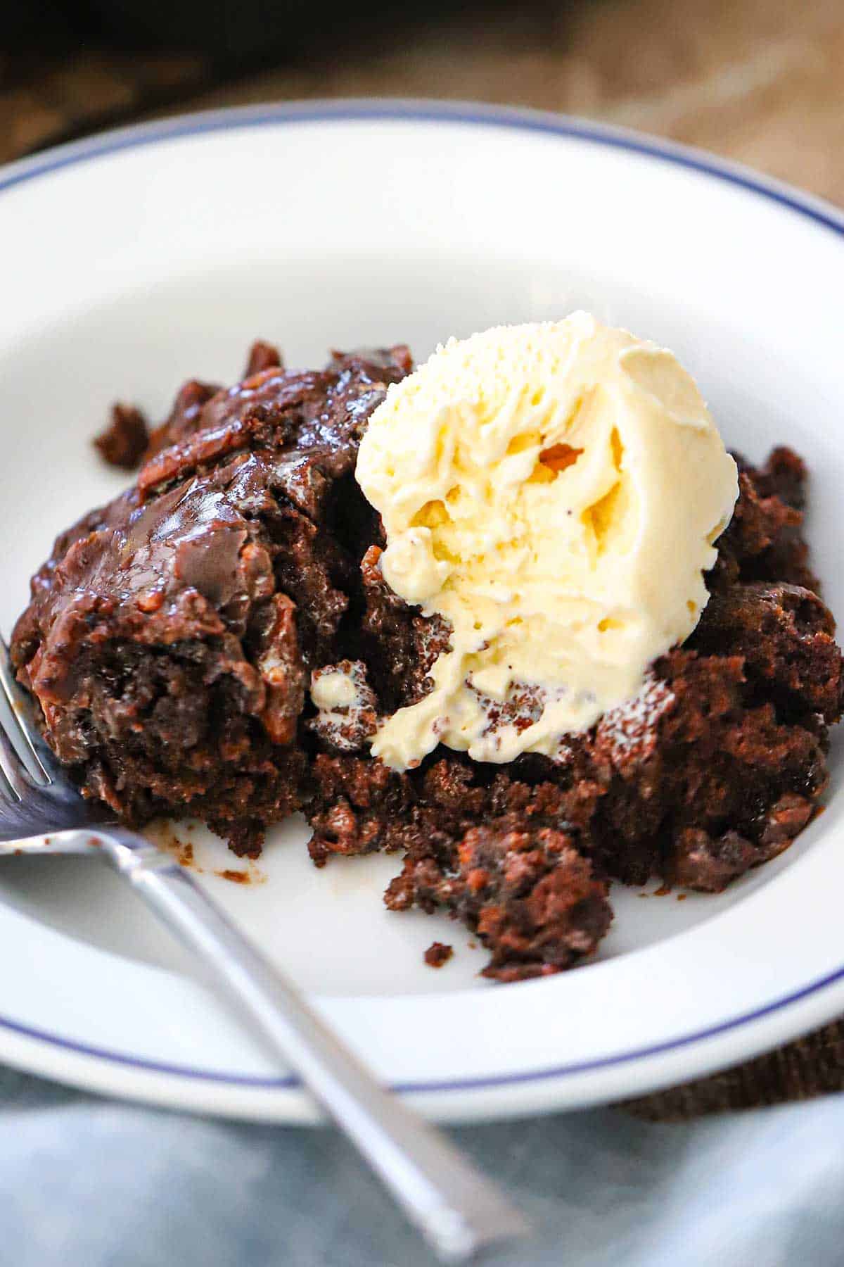 A close-up view of a white bowl filled with a serving of Texas Skillet Cake topped with a scoop of vanilla ice cream that is starting to melt over the cake.