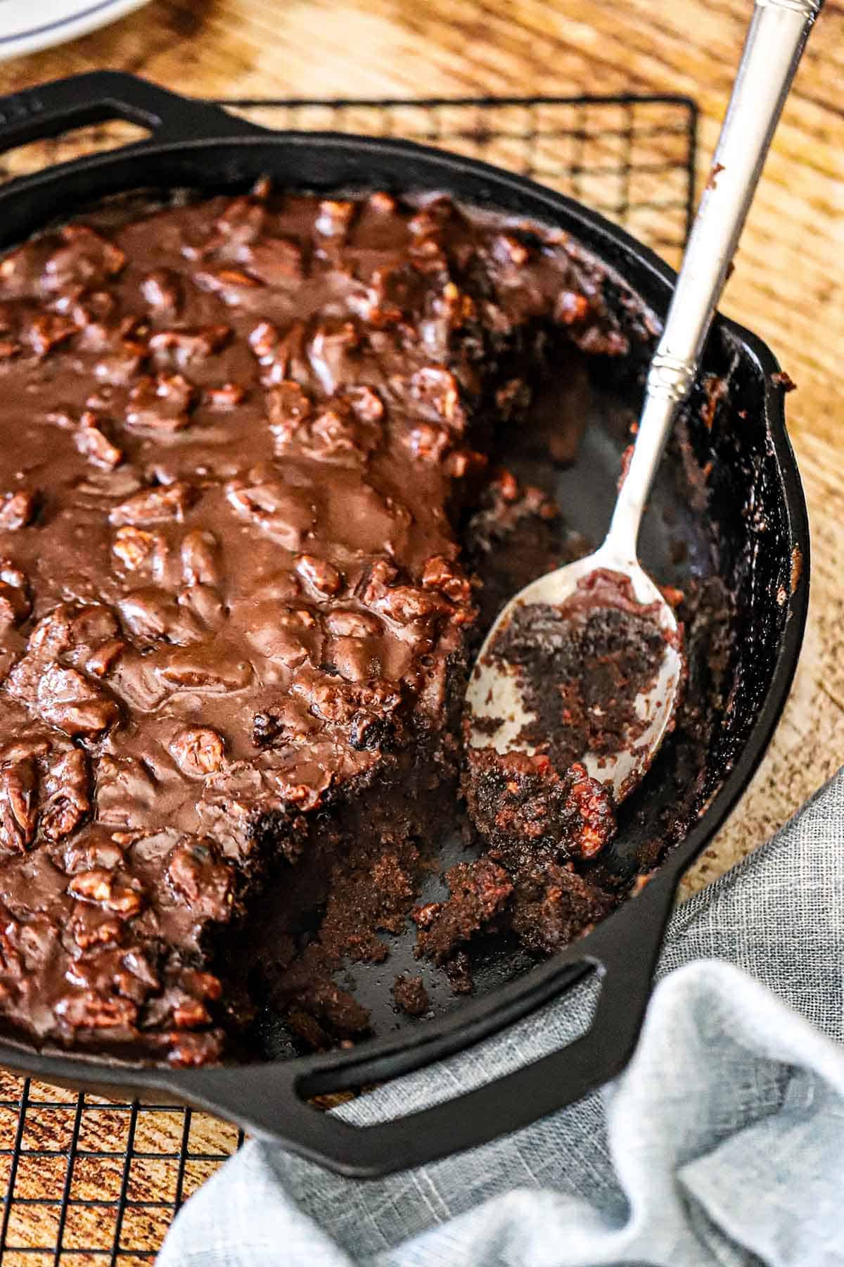 An overhead view of a Fudgy Texas Skillet Cake in a large cast-iron skillet with a portion of it missing and a fudgy serving spoon resting in the pan.