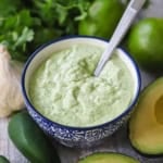 A close-up view of a festive blue and white condiment bowl filled with Jalapeño Lime Sauce with a spoon inserted into it and fresh jalapeños, avocados, and cilantro nearby.