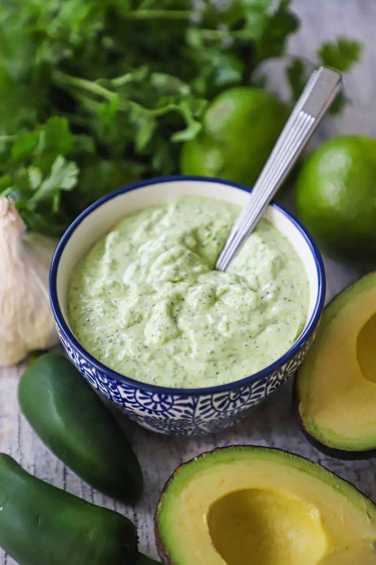 A festive blue and white condiment bowl filled with Jalape&ntilde;o Lime Sauce with a spoon inserted into it and fresh jalape&ntilde;os, avocados, and cilantro nearby.