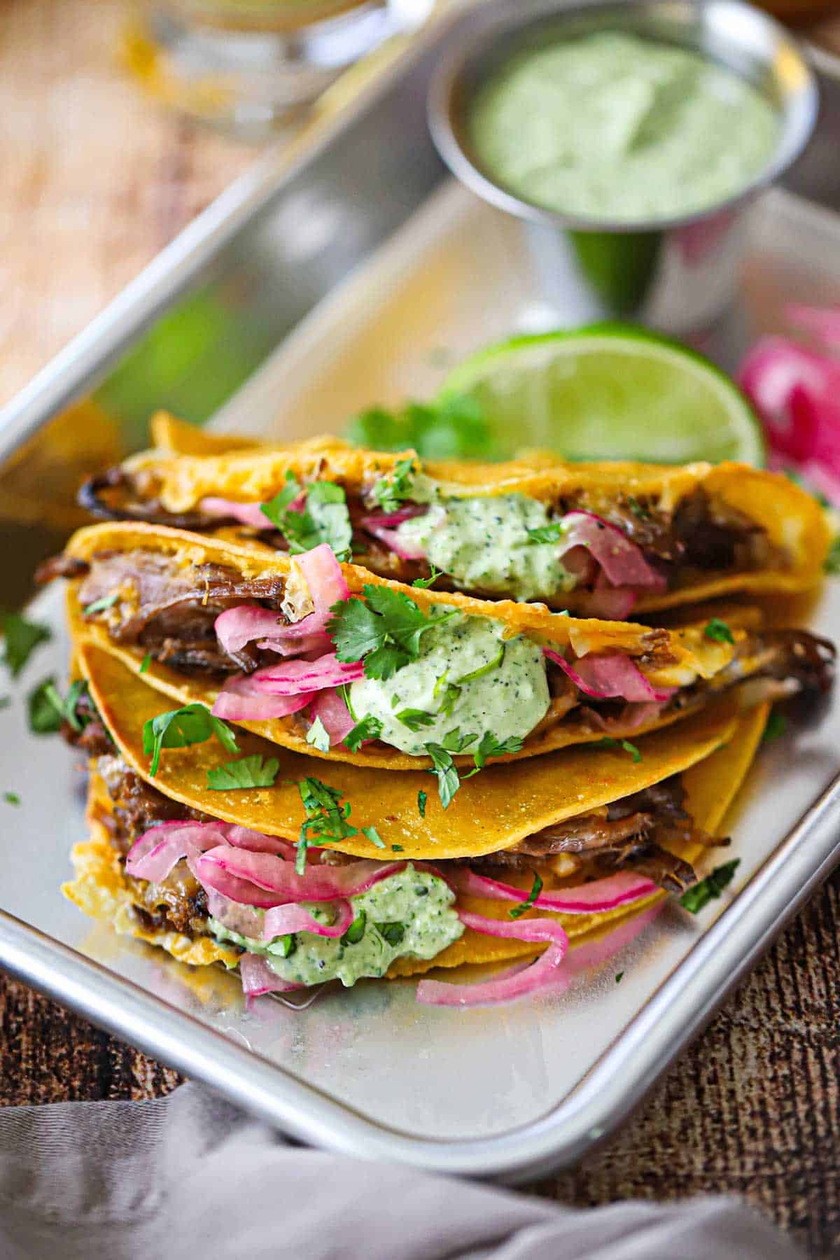 A close-up view of Crispy Baked Brisket Taco garnished with pickled red onions and a dollop of Jalapeño Lime Sauce all on a silver trey.