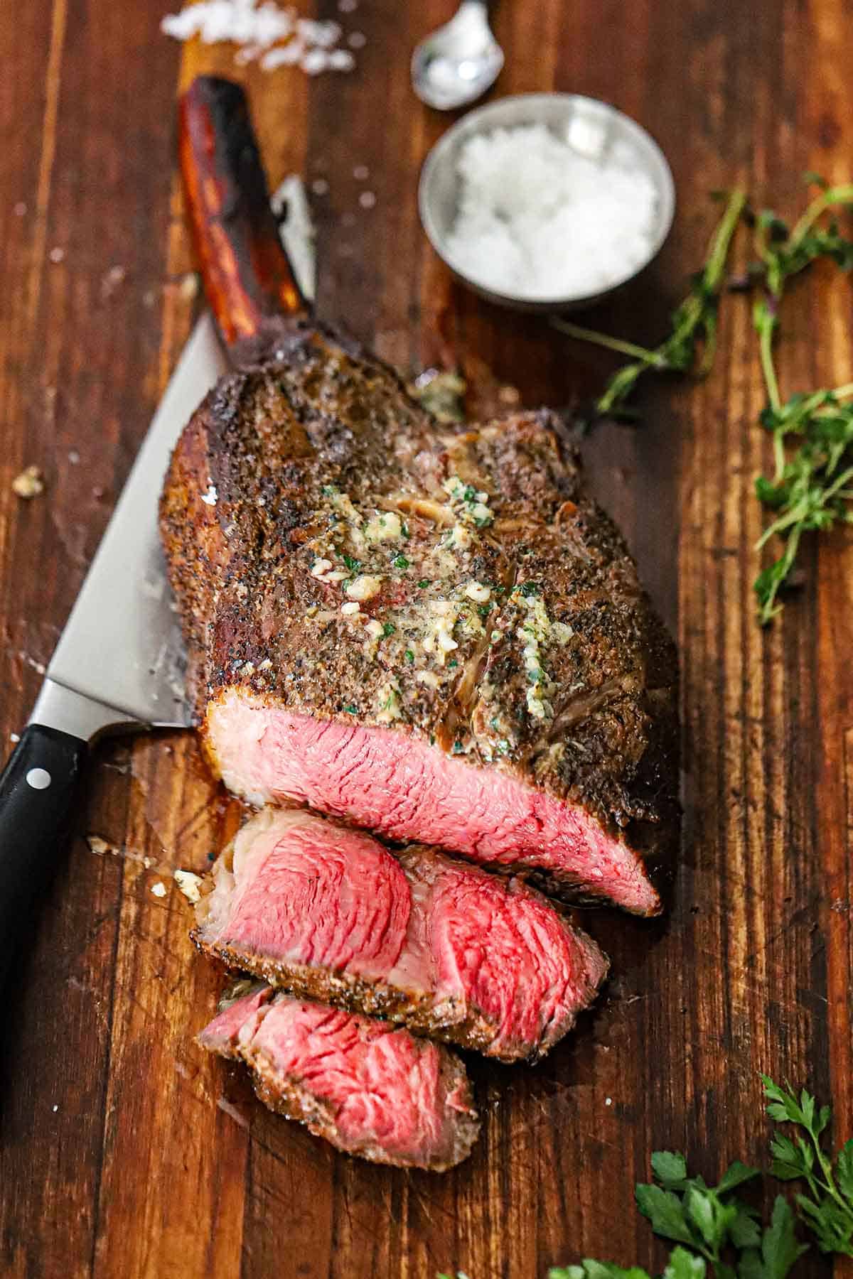 A close-up view of a tomahawk steak with roasted bone marrow butter that has two slices cut from it resting on a wooden cutting board.
