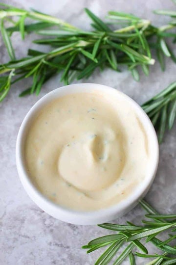 An overhead view of a small white bowl filled with Rosemary Garlic Aioli with several sprigs of rosemary nearby.
