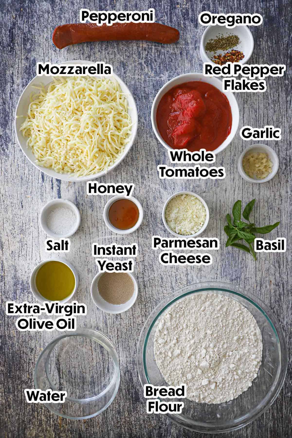 An arrangement of ingredients for sheet pan pepperoni pizza on a grey wooden background including bread flour, water, oil, yeast, honey, garlic, tomatoes, mozzarella, pepperoni, and seasonings.
