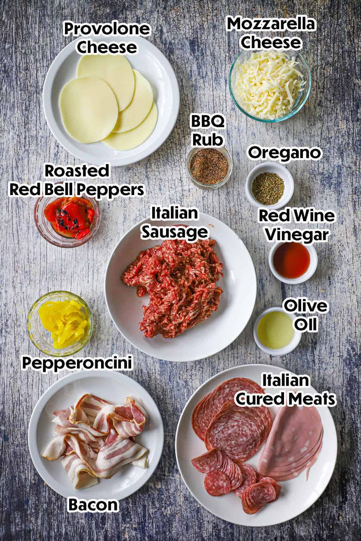 An arrangement of ingredients for a smoked Italian fatty on a wooden background including Italian sausage, Provolone cheese, Mozzarella cheese, roasted peppers, cured meats, bacon, pepperoncini, red wine vinegar, oil, and seasonings.