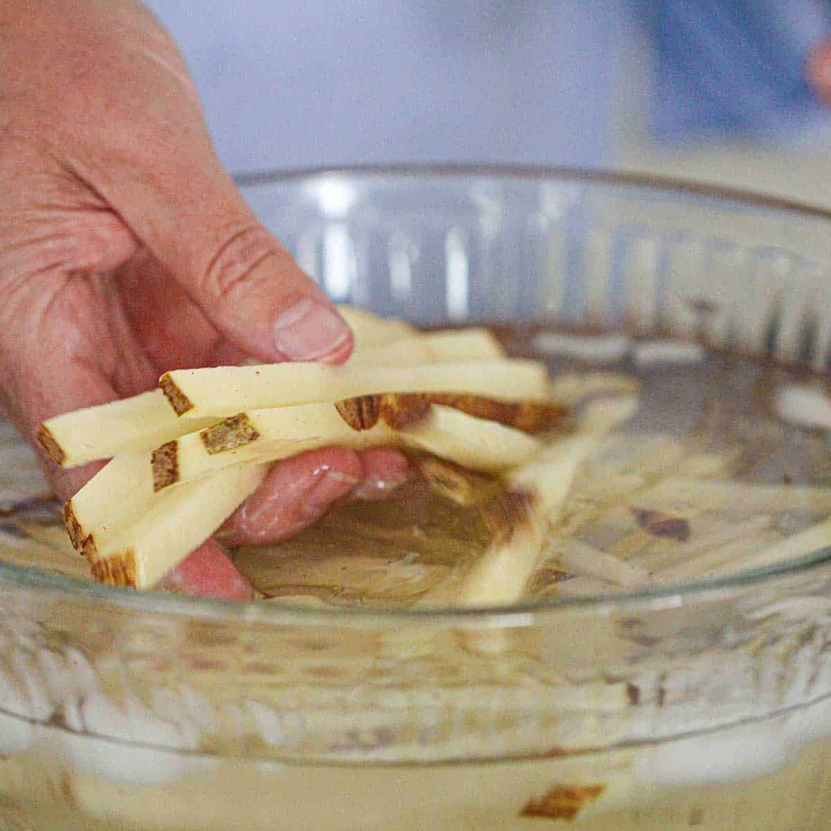 A person swishing julienned strips of Russet potatoes in a large glass bowl filled with water.