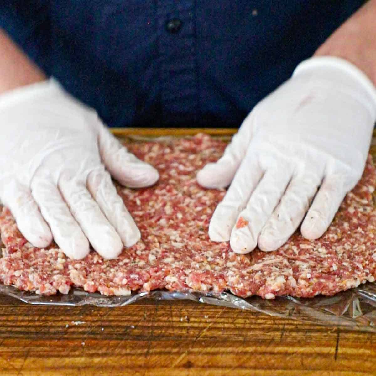A person wearing white latex gloves pressing Italian sausage over the top of a piece of plastic wrap on a wooden cutting board.