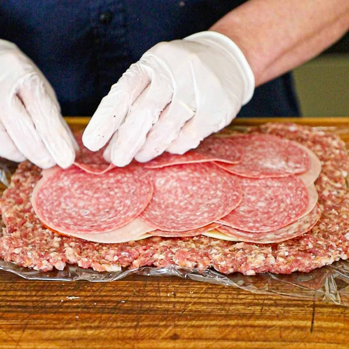 A person placing layers of Italian cold cuts onto the center of a large rectangular shape of flattened Italian sausage onto of a piece of plastic wrap.