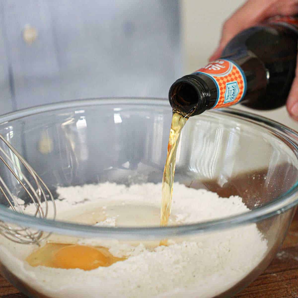 A person pouring beef from a bottle into a glass bowl filled with seasoned flour and an egg.