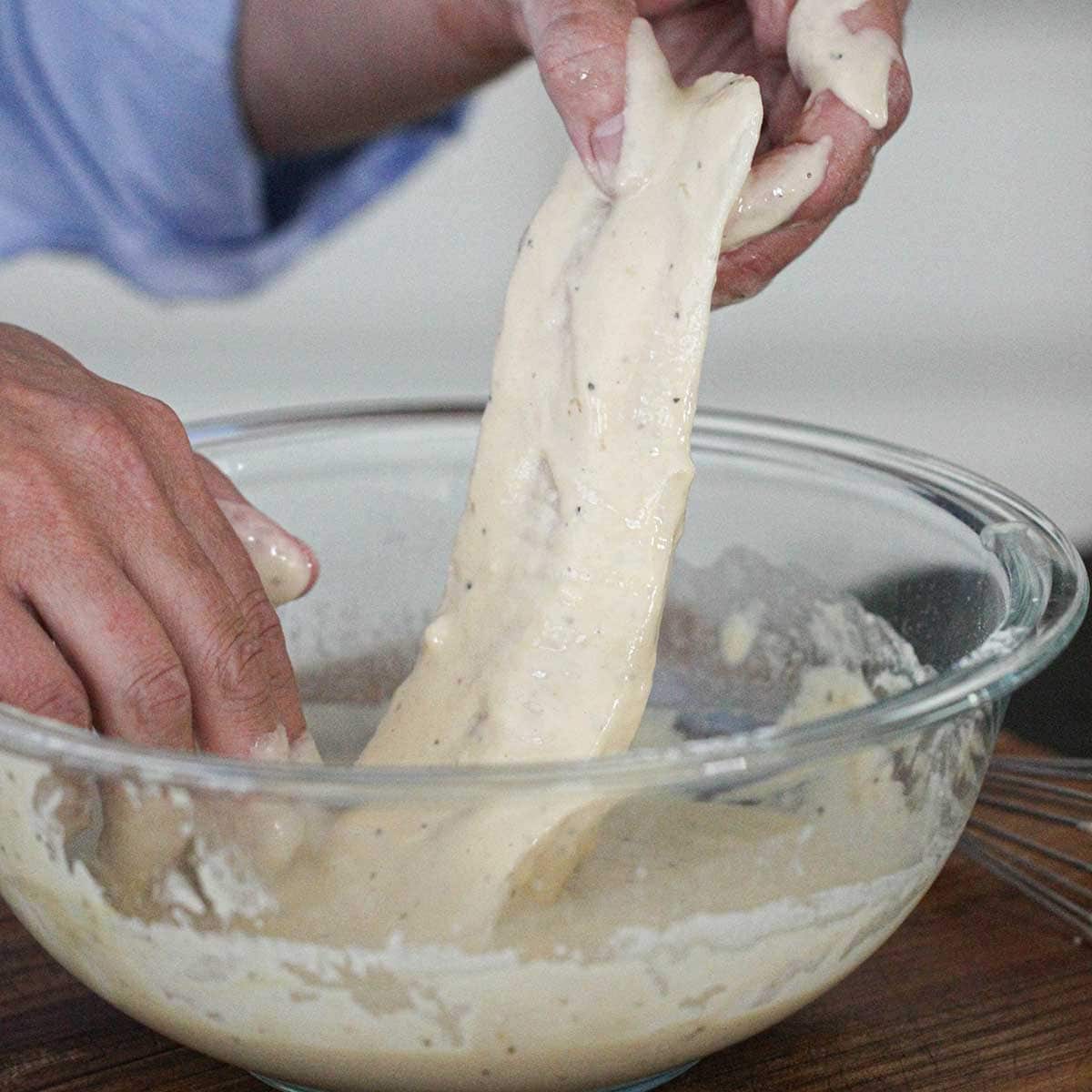 A person holding a filet of cod that had submerge in a wet fish and chips batter in a large glass bowl.