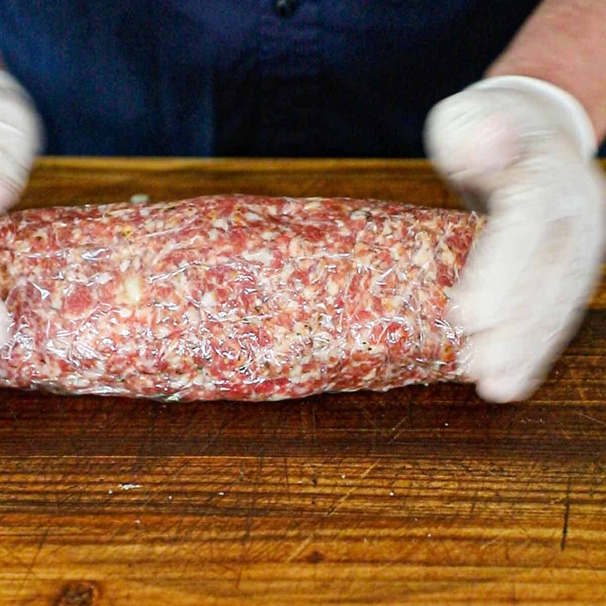 A person using his hand to compact a tightly rolled Italian fatty in plastic wrap on a wooden cutting board.