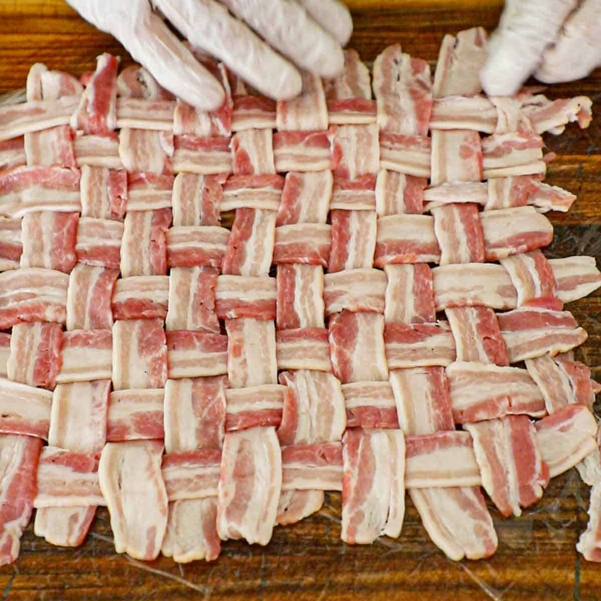 A person using his hands to weave strips of bacon together to form a large braided square on a wooden cutting board.