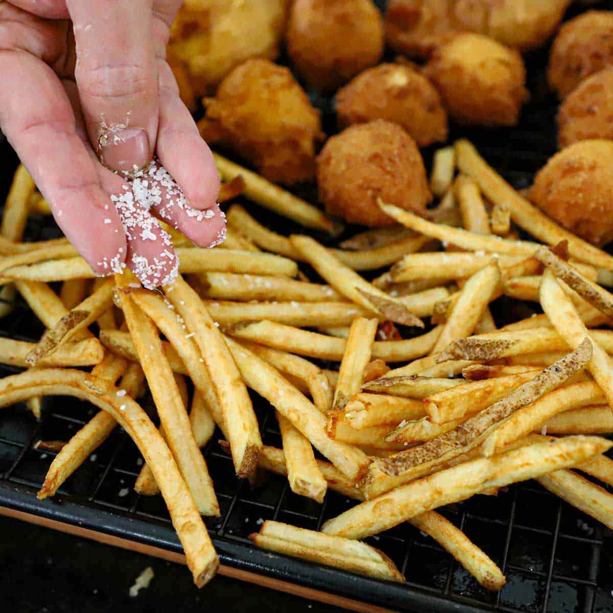 A person using his fingers to sprinkle coarse sea salt over homemade French fries on a baking rack next to homemade hush puppies.