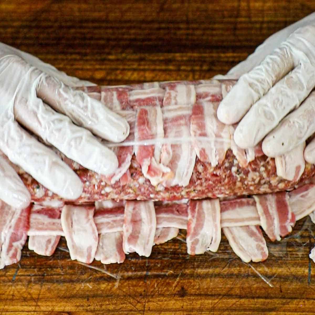 A person using plastic wrap to encase an uncooked Italian fatty in a breaded square of bacon.