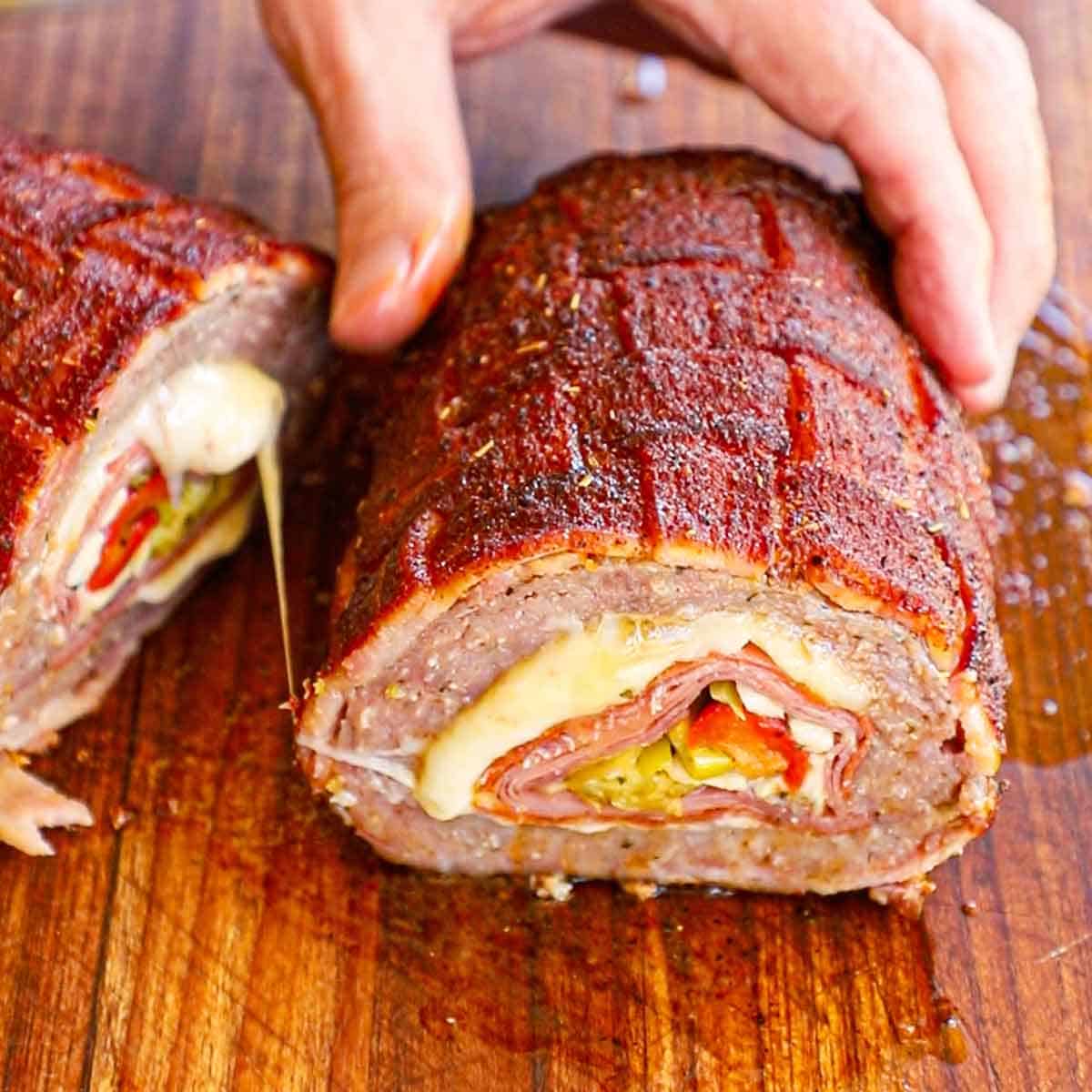 A person holding one half of a Smoked Italian Fatty revealing the fully cooked Italian meats, melty cheese, and roasted veggies inside.