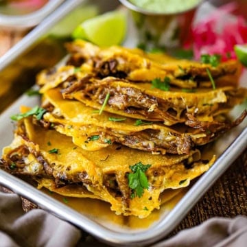 A close-up view of crispy oven baked brisket tacos on a small metal trey with a lime wedge and a small pile of pickled red onions in the rear or the pan.