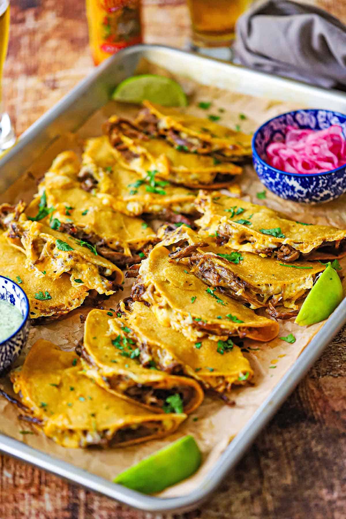 A sheet pan filled with two rows of baked brisket tacos with a colorful bowl filled with pickled red onions in one corner and another bowl filled with jalapeño-lime sauce in the opposite corner.