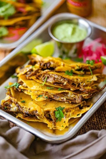 A row of four crispy oven baked brisket tacos on a small metal trey with a lime wedge and a small pile of pickled red onions in the rear or the pan.