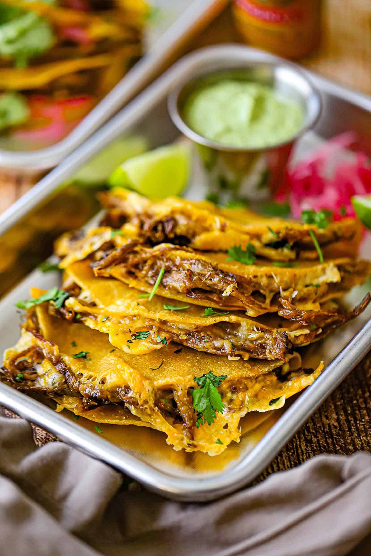 A row of four crispy oven baked brisket tacos on a small metal trey with a lime wedge and a small pile of pickled red onions in the rear or the pan.
