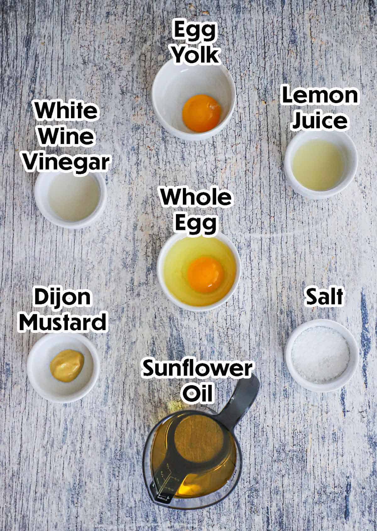 An arrangement of ingredients for homemade mayonnaise on a grey wooden background including an egg yolk, a whole egg, vinegar, lemon juice, Dijon mustard, salt, and oil.