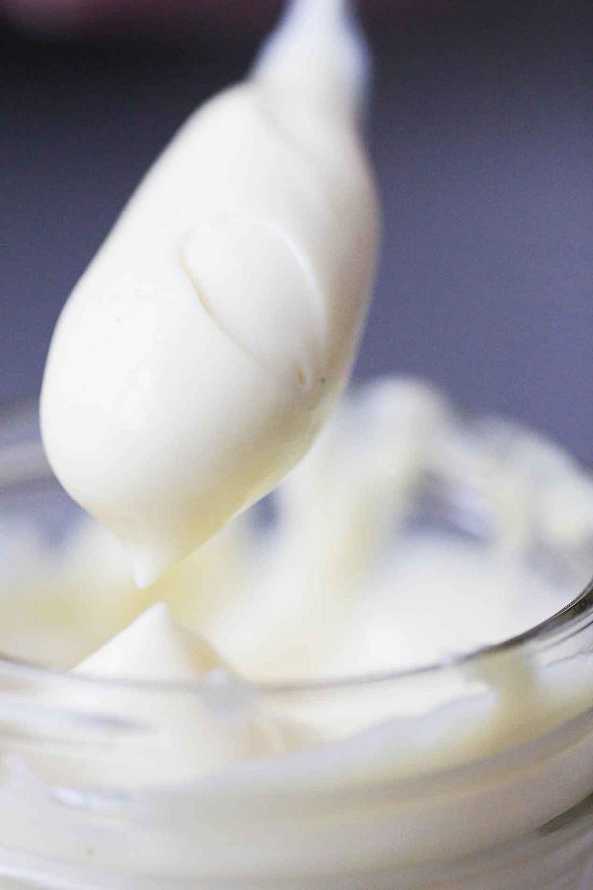 A person holding a spoon the is completely covered with a dollop of homemade mayonnaise above a glass jar filled with the same.