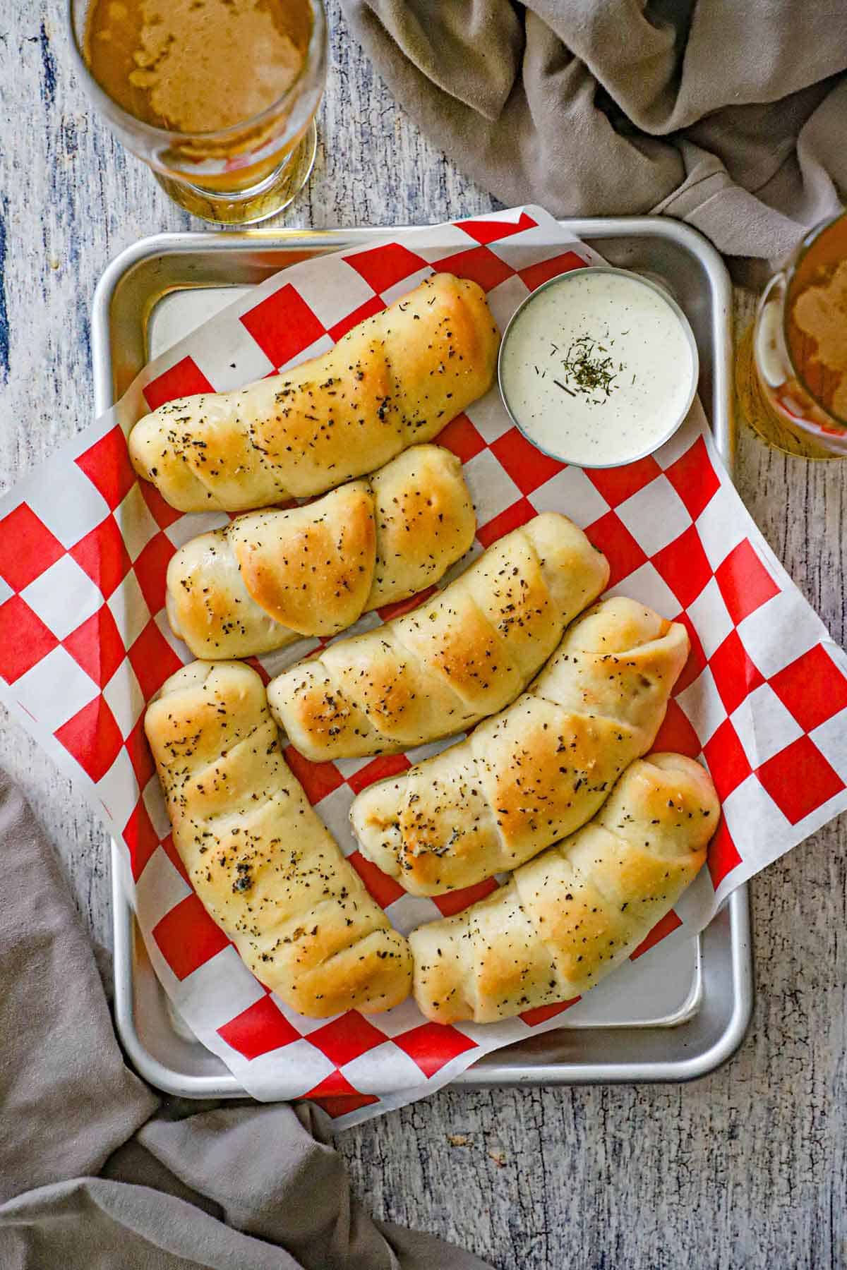 An overhead view of homemade Melt-In-Your-Mouth Pepperoni Rolls resting in metal trey lined with a red checkered piece of wax paper and a bowl of Ranch dressing up in the corner.
