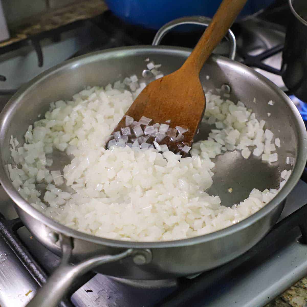 A person using a wooden spatula to stir a large amount of chopped onions that are being sautéed in a large skillet on a gas stove.