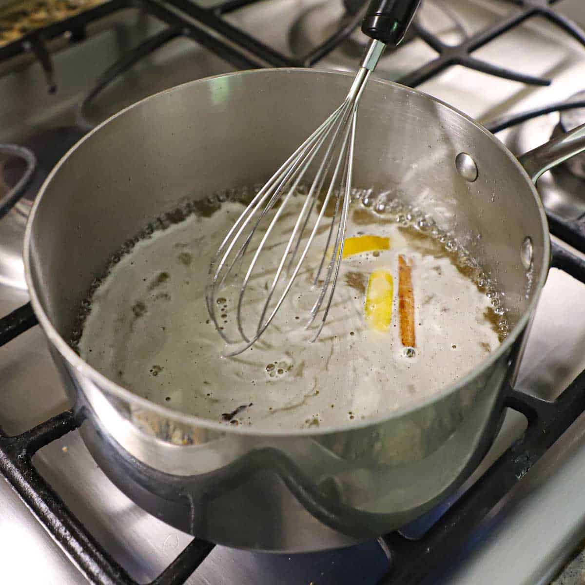 A person using a wire whisk to stir simmering sugar water with honey, a cinnamon stick, lemon peel, and whole cloves.