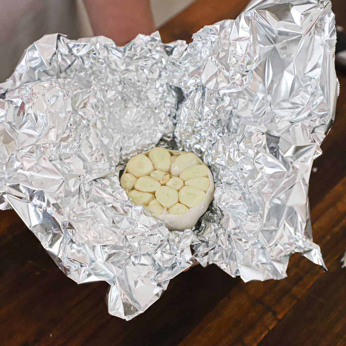 A person holding a piece of aluminum foil that is being wrapped around a head of garlic that has had the end of it cut off revealing the cloves of garlic inside.