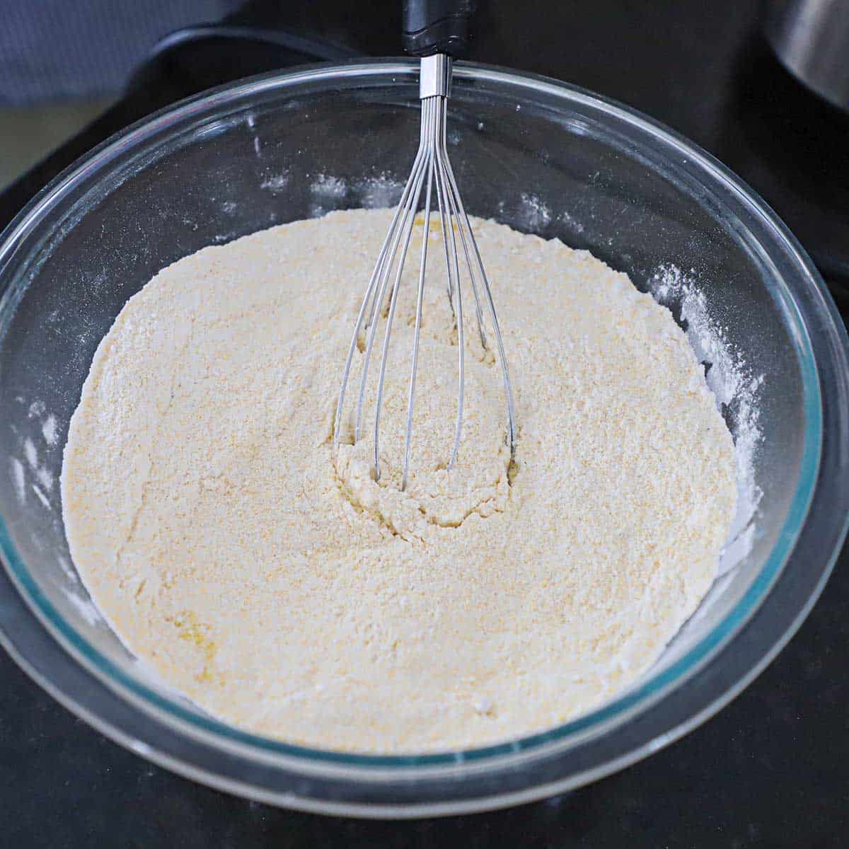 A person using a whisk to combine flour, cornmeal, sugar, baking and powder in a large glass bowl.