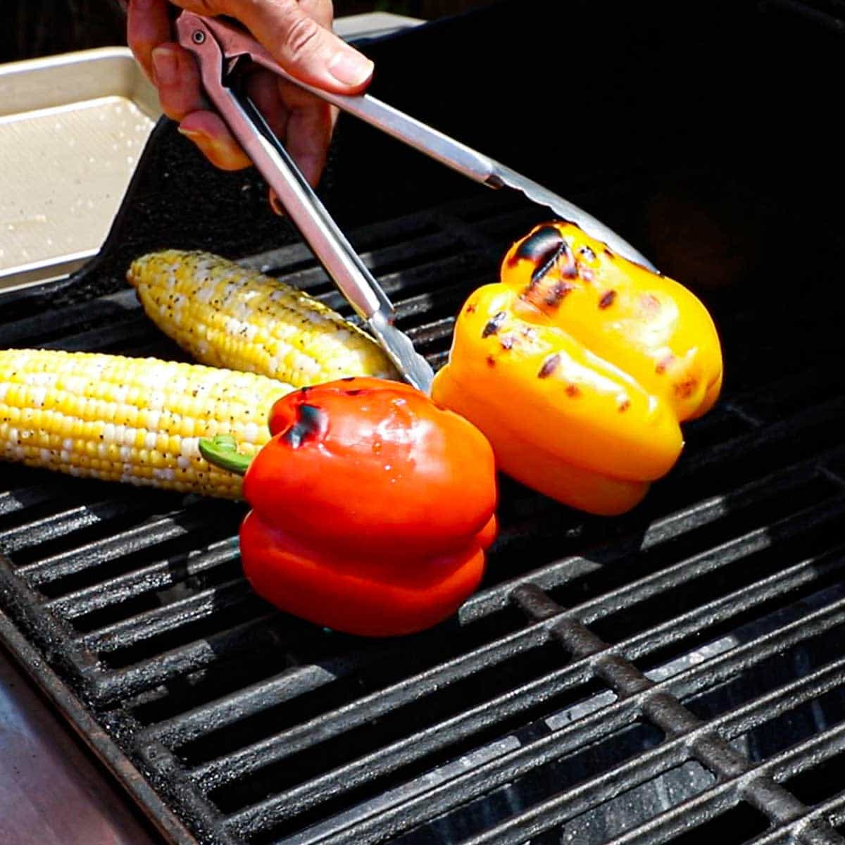A person using a pair of metal tongs to turn and roast bell peppers on a gas grill along with two ears of corn.
