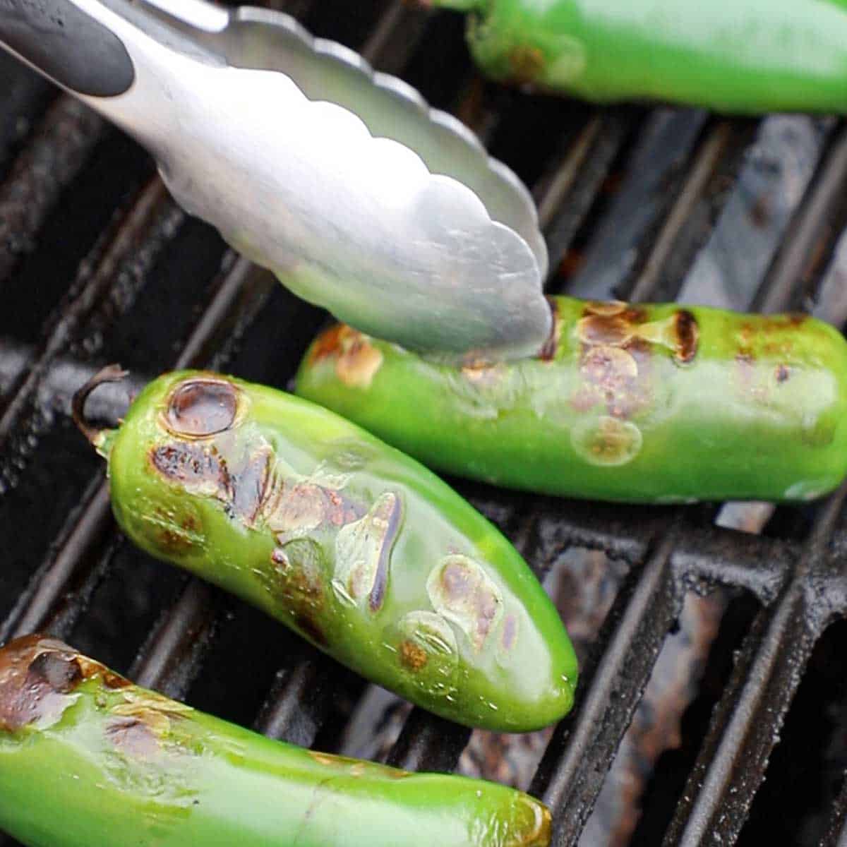 A person using a pair of metal tongs to turn fresh jalapeños that are being roasted on the grate of a gas grill.