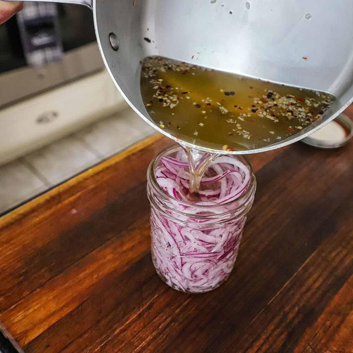 A person pouring a pickling broth into a glass jar that is stuffed and filled with thinly sliced red onion.