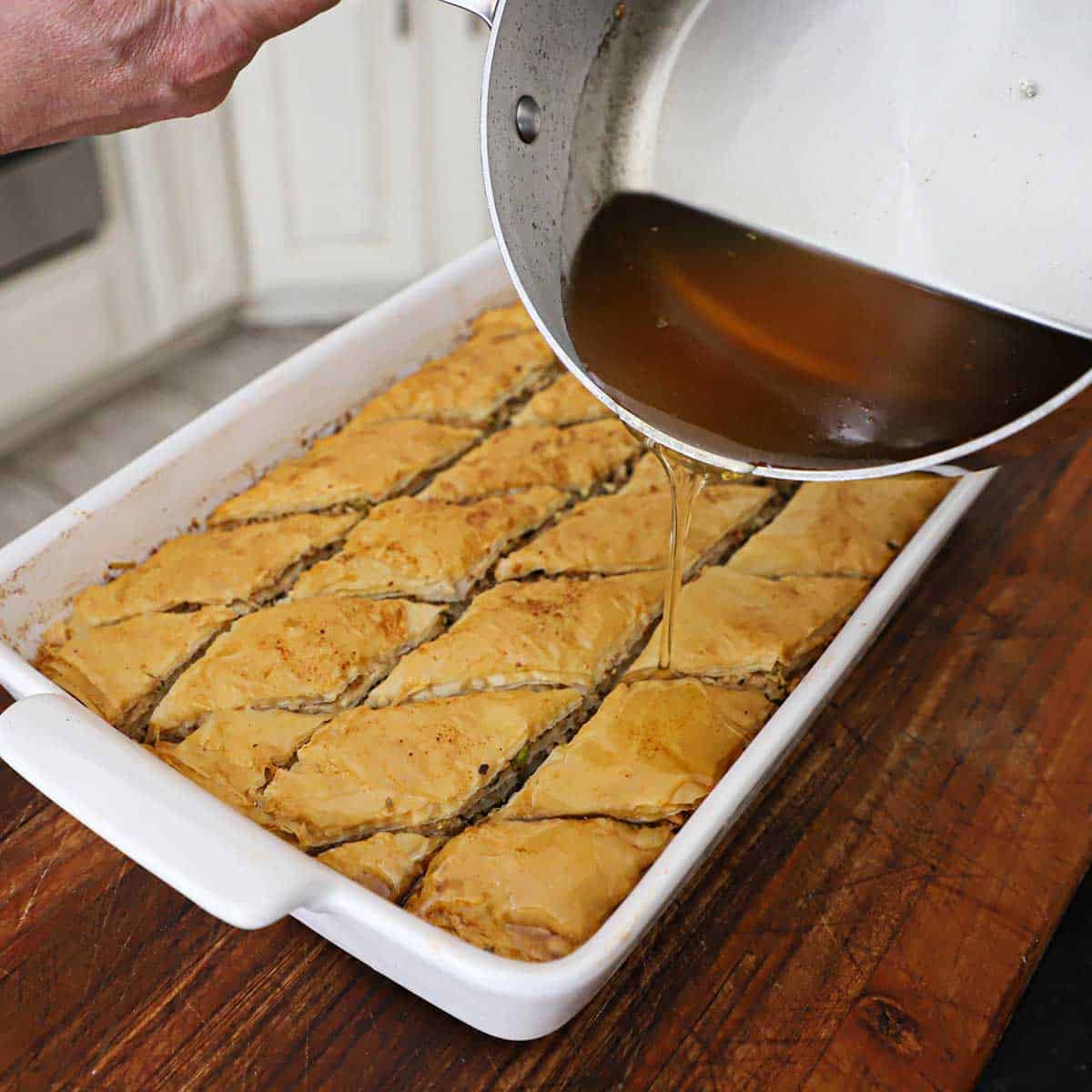 A person pouring a honey sugar glaze from a saucepan over the top of a freshly baked dish of Authentic Greek Baklava.