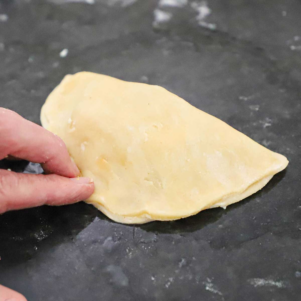A person using his fingers to press the edges of a folded over uncooked Argentine Beef Empanada on a black marble counter.