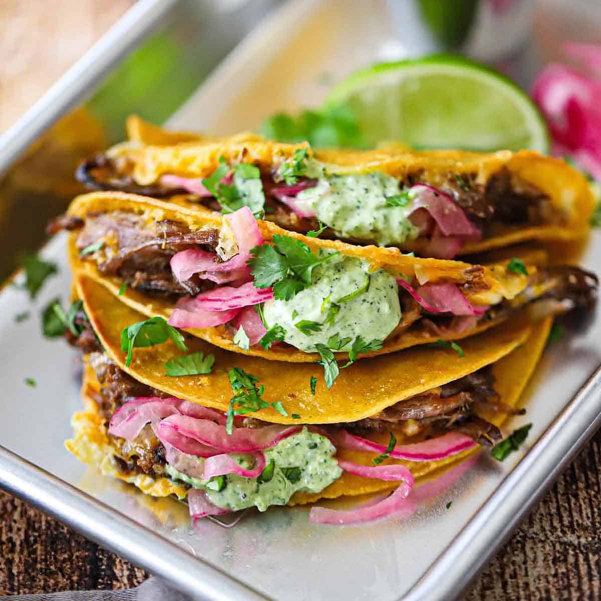 A close-up view of crispy baked brisket tacos that have been garnished with pickled red onions, a dollop of jalapeño-lime sauce, and chopped cilantro.