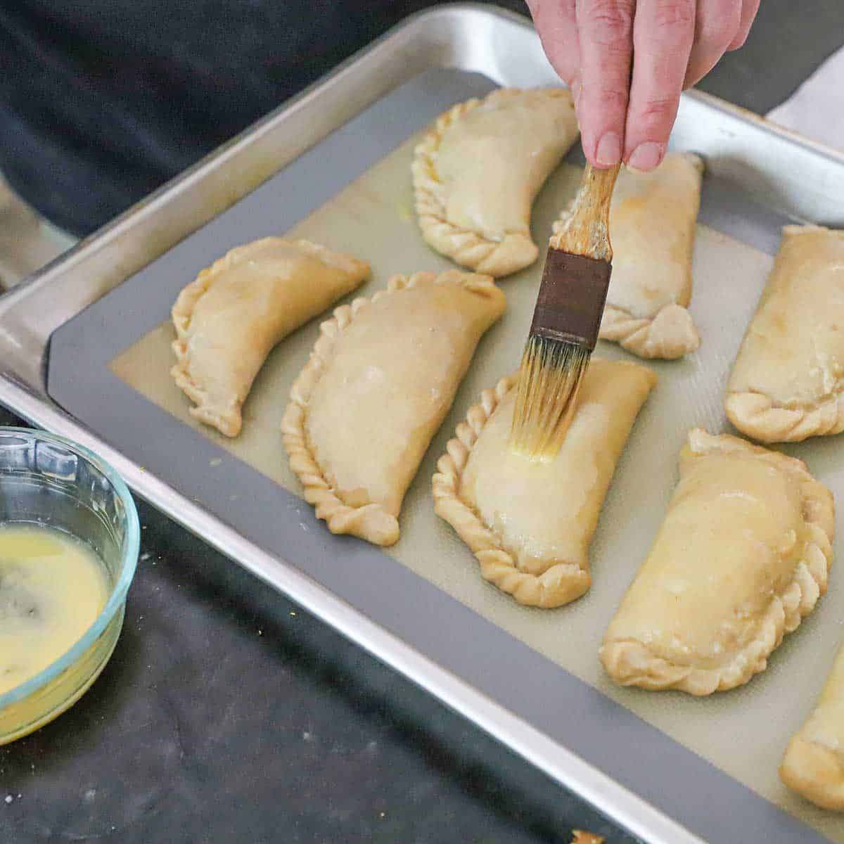 A person using a pastry brush to apply an egg wash over the tops of uncooked beef empanadas on a baking pan lined with a silicone mat.