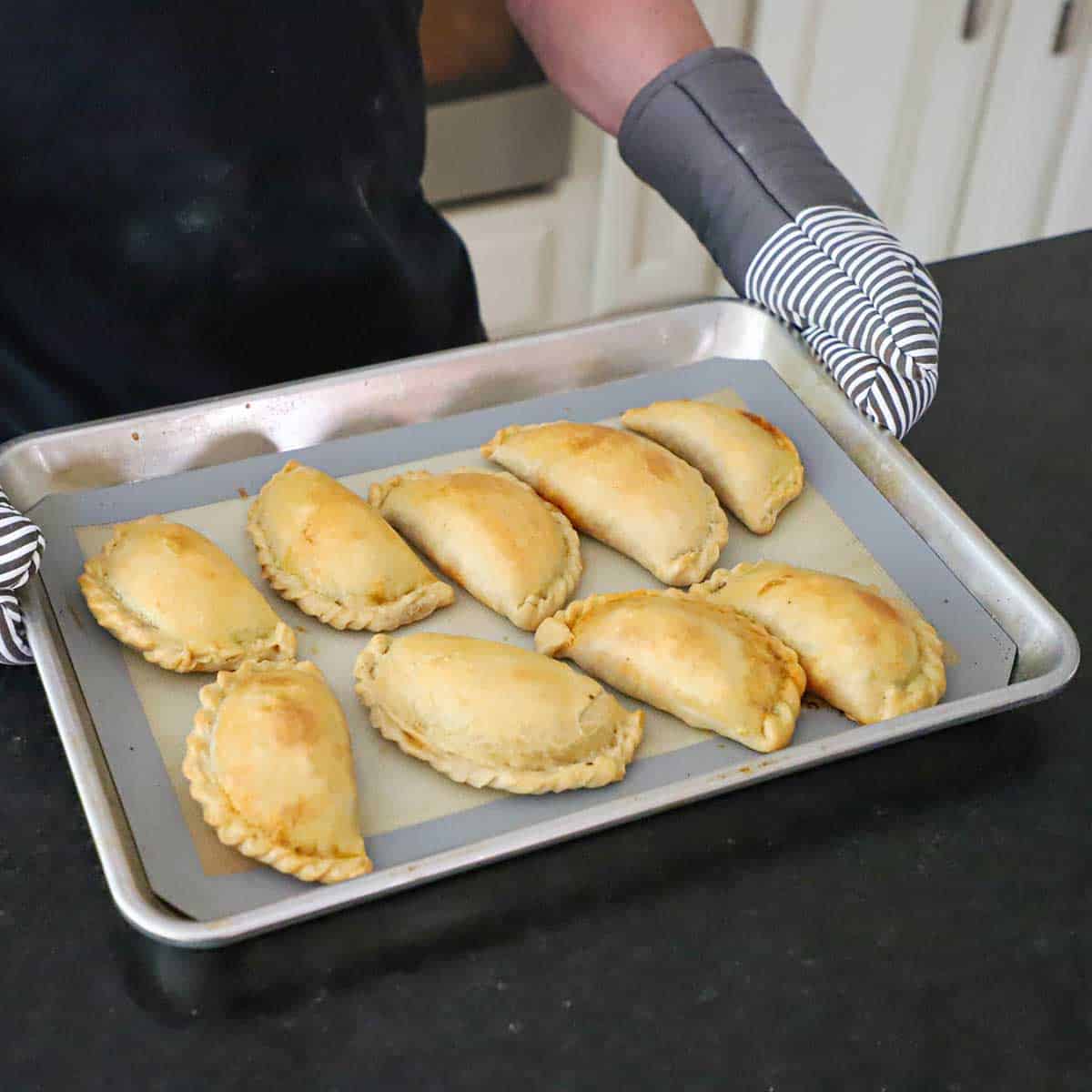 A person with oven mitts holding a baking pan that is filled with freshly baked Argentine Beef Empanadas.