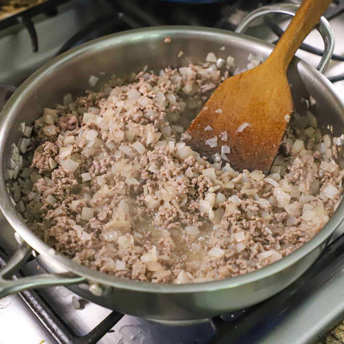 A person using a wooden spatula to stir ground beef and chopped onions that are being cooked in a large skillet on a gas stove.