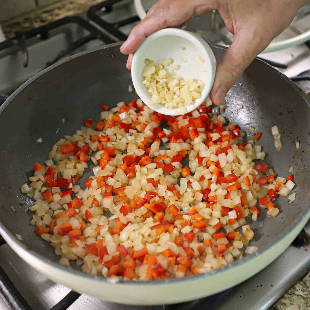 A person transferring minced garlic from a small bowl into a large skillet filled with saut&eacute;ed chopped onions and red bell pepper.