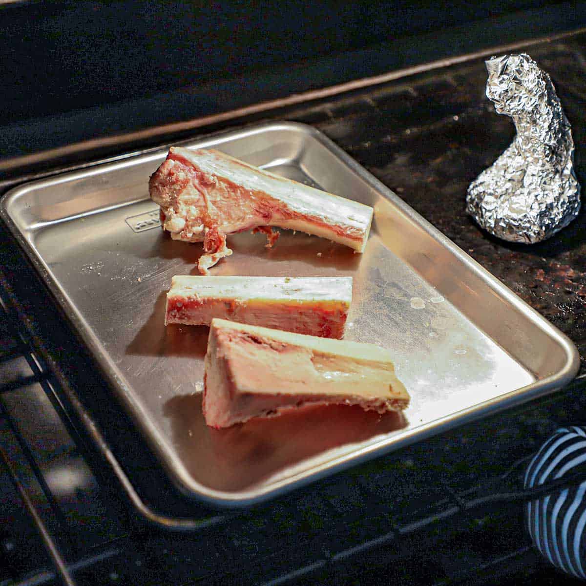 A person placing a small sheet pan into an oven with three beef bone marrow canoes on it and a wrapped piece of foil filled with garlic sitting next to the pan.