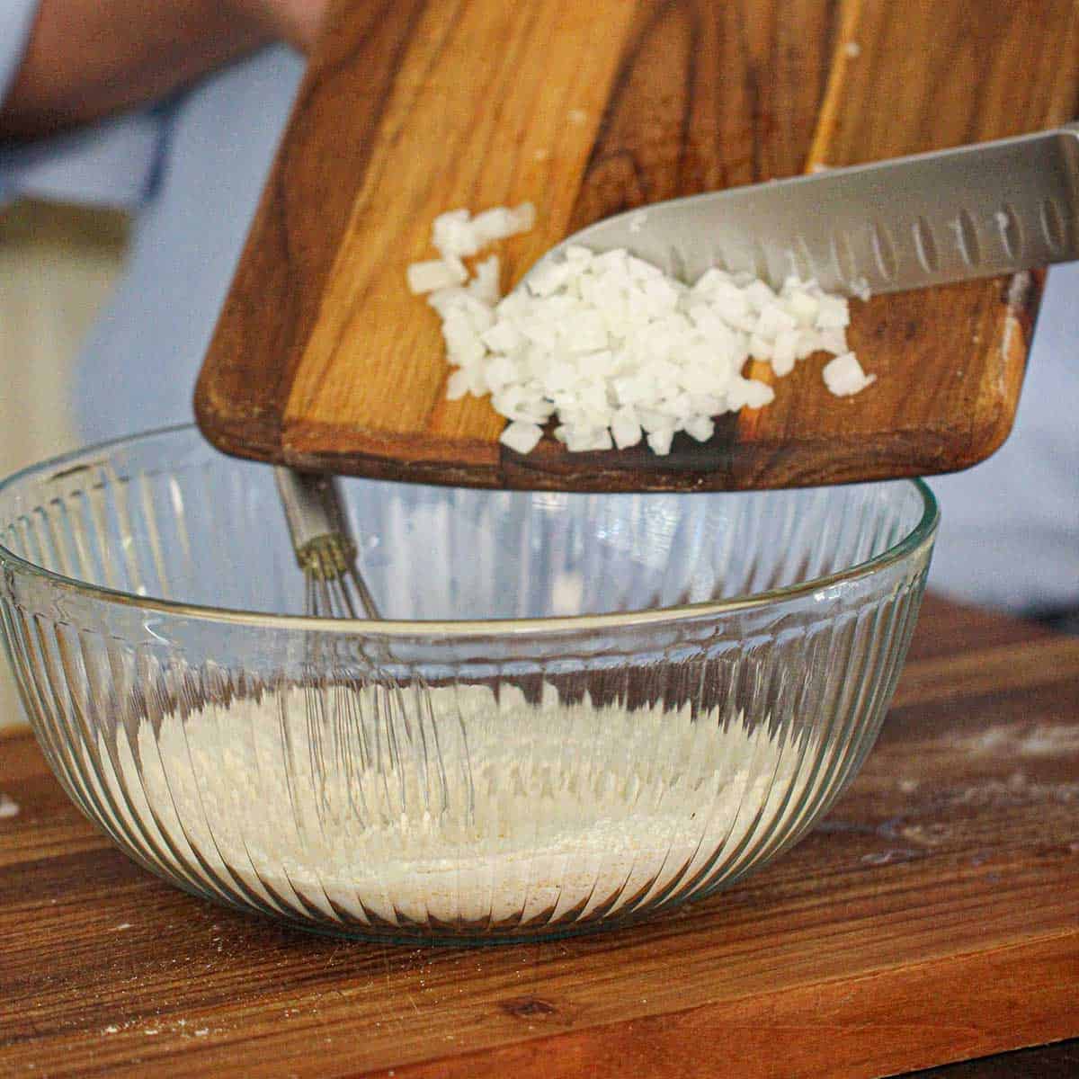 A person transferring chopped into a large glass bowl filled with the dry ingredients for Southern Hush Puppies.