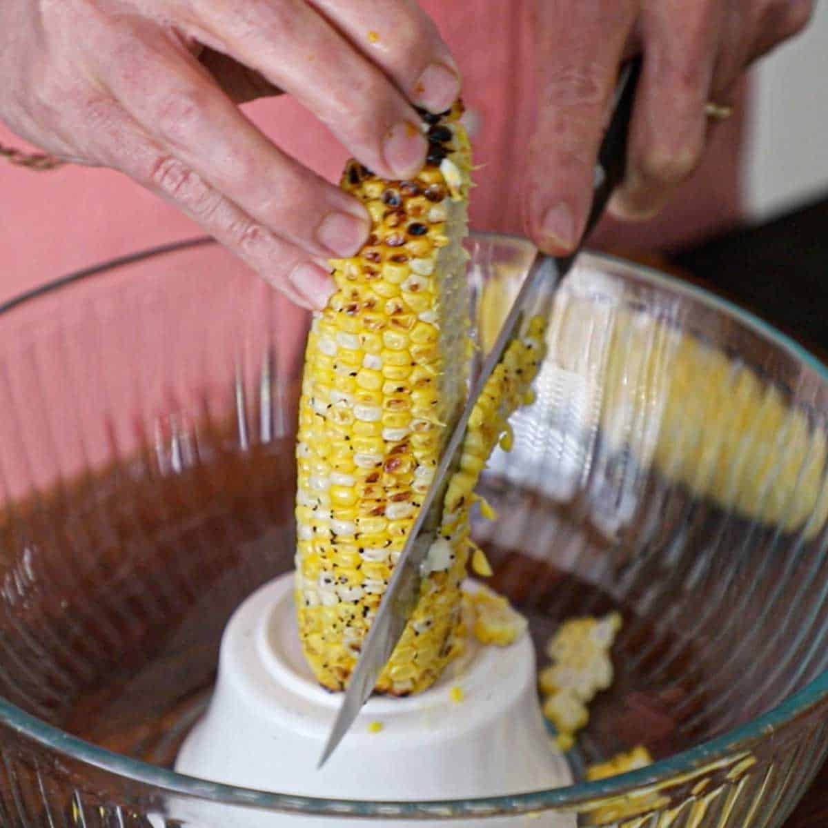 A person using a chef's knife to cut grilled corn kernels from a cob that is resting on a small inverted bowl inside a large glass bowl.